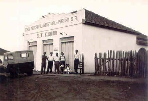 Foto antiga da fachada de uma agência do Banco Mercantil e Industrial do Paraná, com funcionários na porta e um jipe estacionado